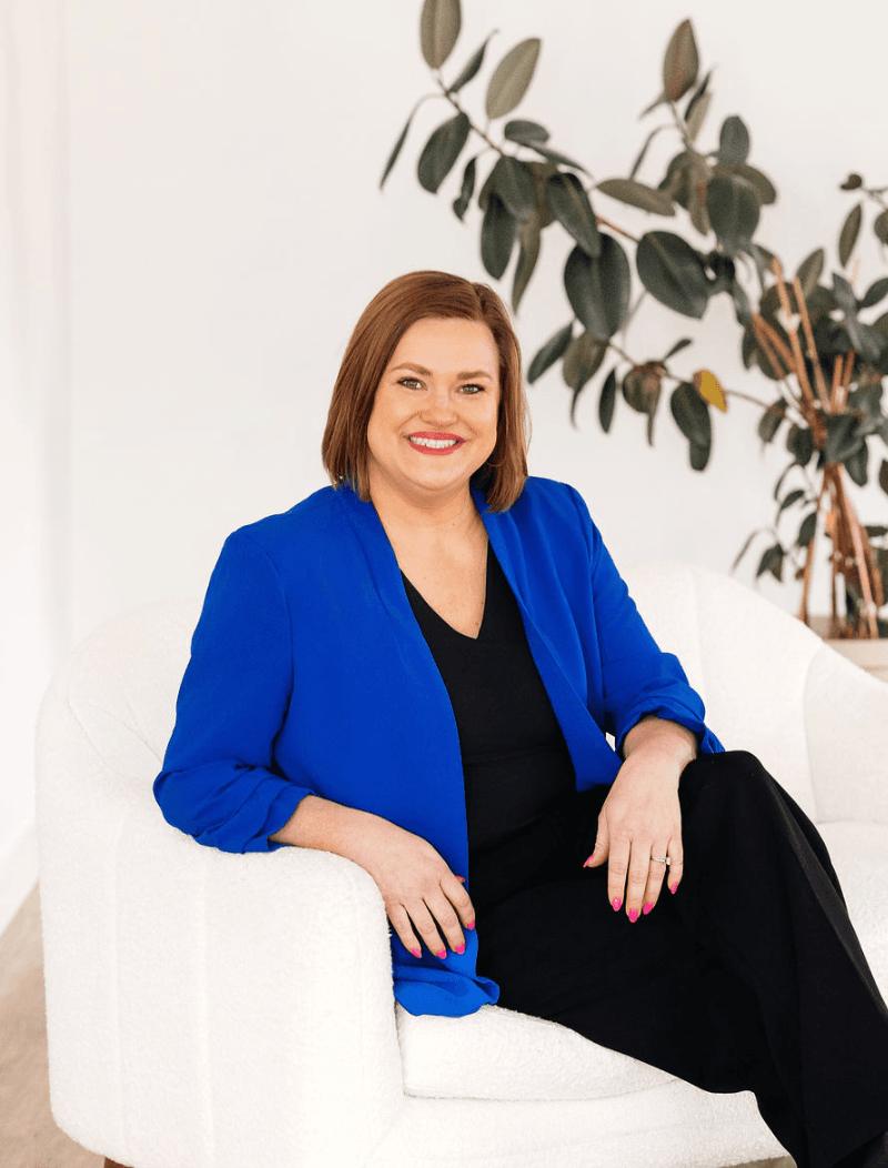 Woman in a blue blazer sitting on a white chair in front of a plant.