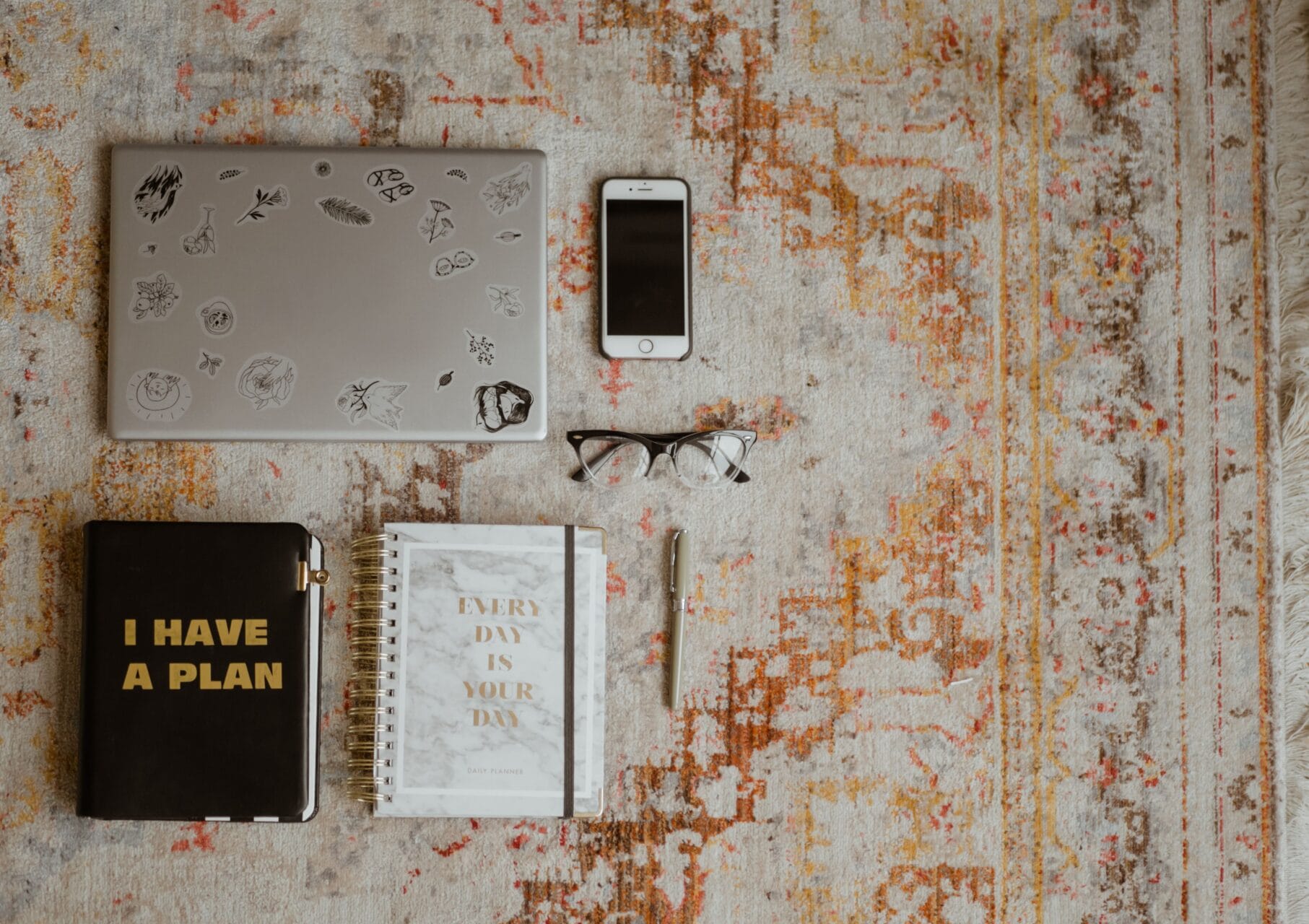 A desk with a laptop, cell phone, glasses and two notebooks and a pen.