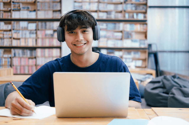 Young man wearing headphones, sitting in front of a book shelf with a laptop.