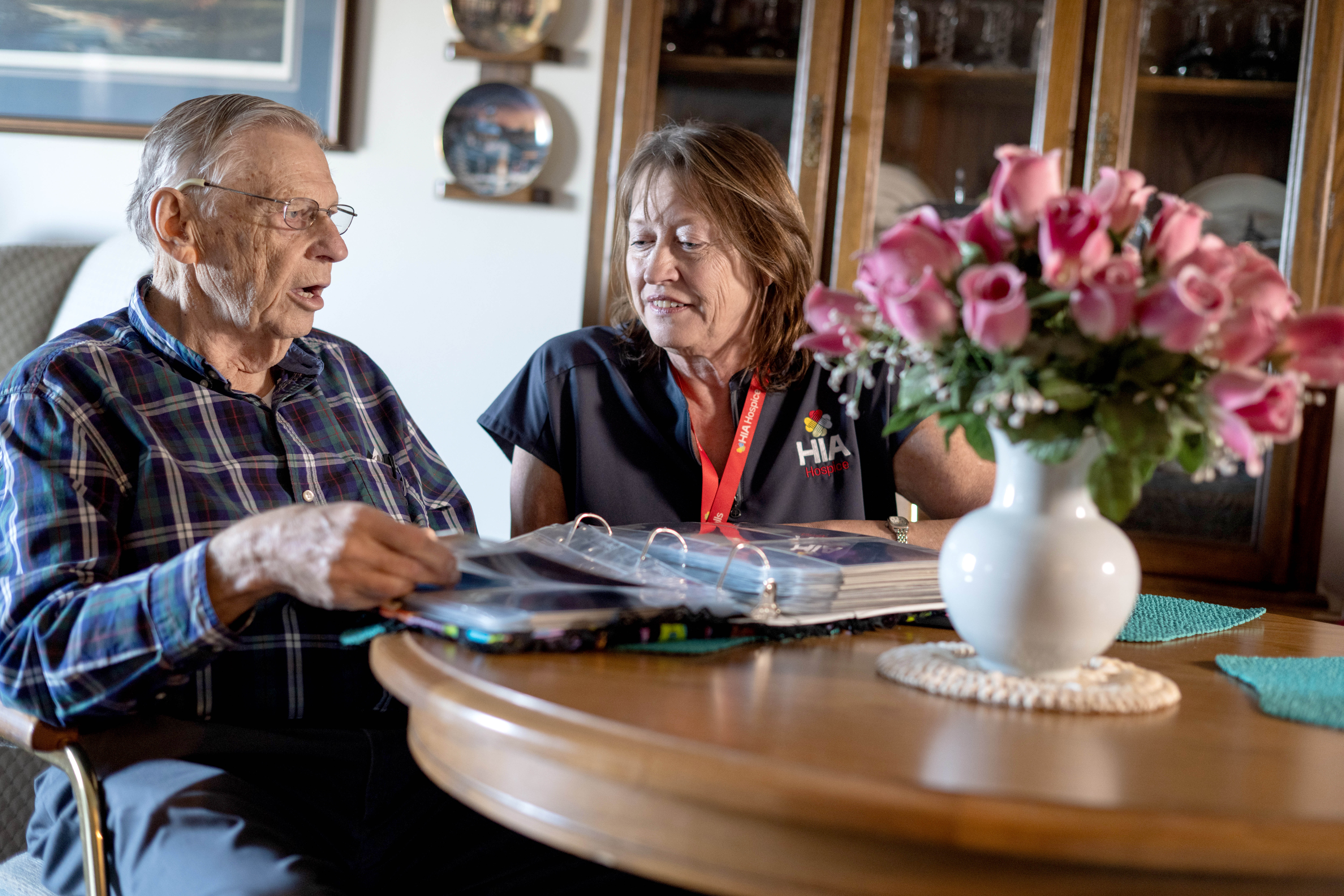 An elderly man sitting at a table with a Hospice worker. There is a white vase with pink flowers on the table.